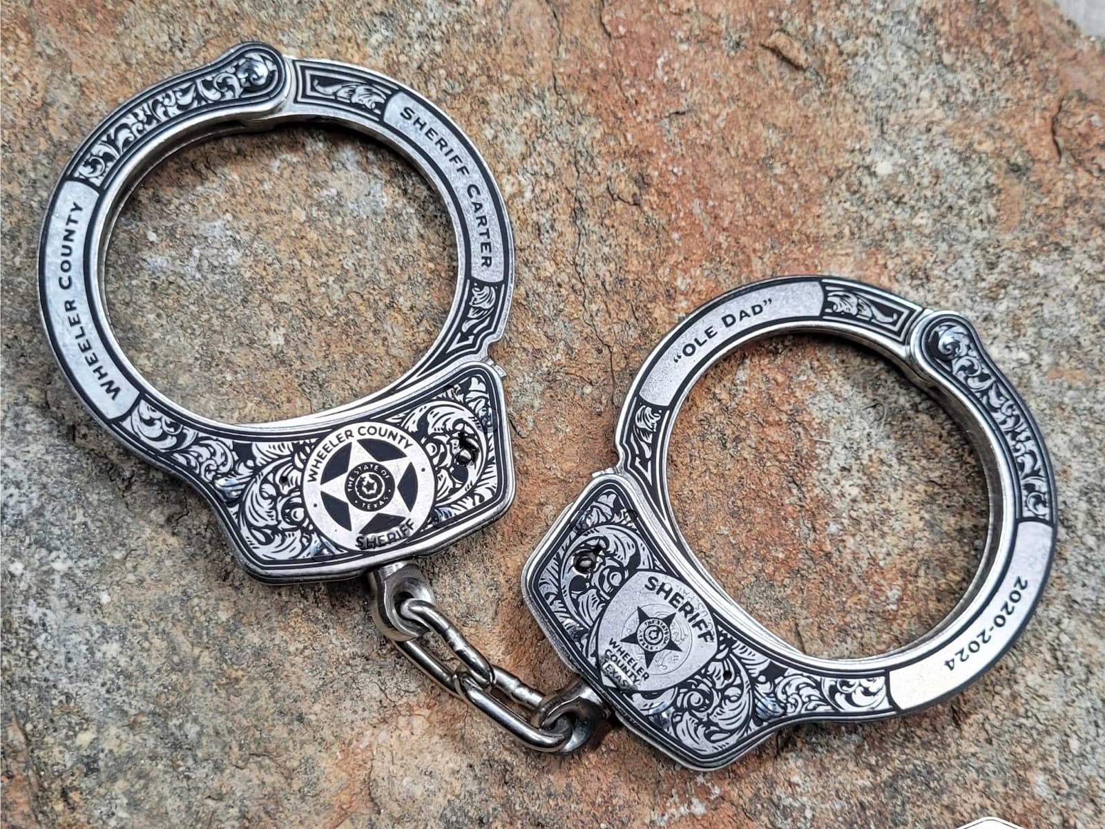 A pair of nickel handcuffs engraved in the Double Scroll design, resting on stone with a star badge on each plate and a connecting chain.
